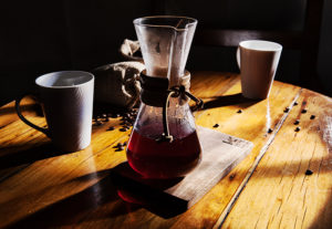 A low-key moody photo of a Chemex coffee jug bathed in partial bay window light. Surrounded by scattered coffee beans and two white mugs.