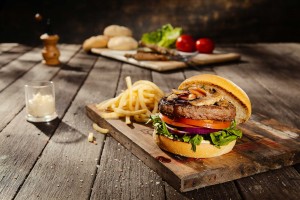 A hamburger with french fries on a wood table in dappled outdoor light with food out of focus in the background