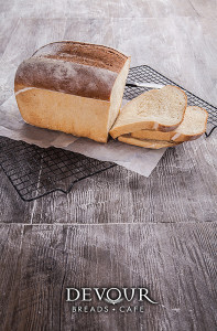 An image of a loaf of artisanal white bread presented on baking paper and metal tray, atop a timber tabletop, surrounded by scattered flour.