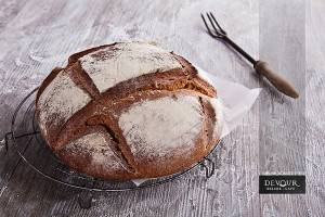 An image of a round loaf of artisanal crusty bread with a rustic serving fork in the background, on a timber tabletop, surrounded by scattered flour.