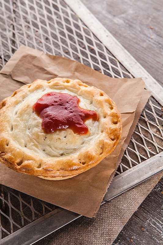 An image of a pie with tomato sauce sitting on a brown paper bag and tin baking trays on a timber tabletop.