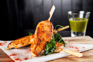 A photo of a chicken skewers served on a wooden food board, sprinked with seasoning powder with a soft focus glass of satay in the background
