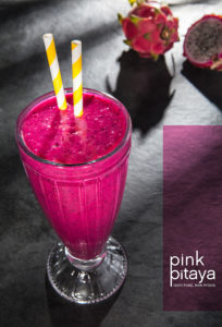 A photo of a tall Pink Pitaya (Dragonfruit) smoothie glass on a black slate table with a raw uncut Pitaya (Dragonfruit) in the background