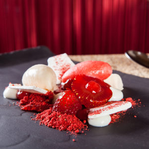A photo of an elaborately-presented fine dining dessert served on a black earthen platter with rich red drapery in the background