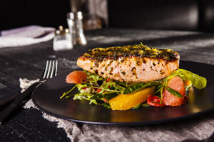 An image of a cooked salmon fillet on a black rustic timber restaurant table