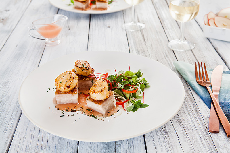 A fine dining dish of scallops atop pork belly squares, presented beautifully on a white plate with garden salad.